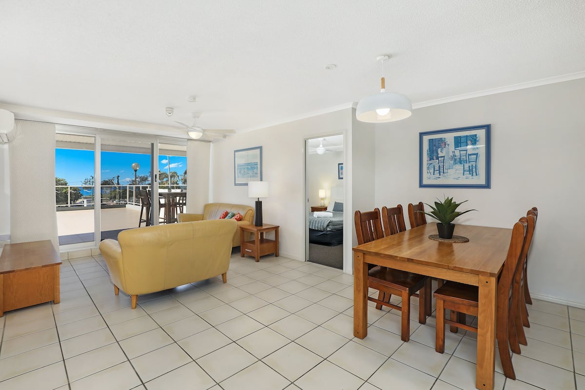 A spacious living area with a yellow sofa, wooden dining table, and chairs. There is a balcony with a view, and a ceiling fan.