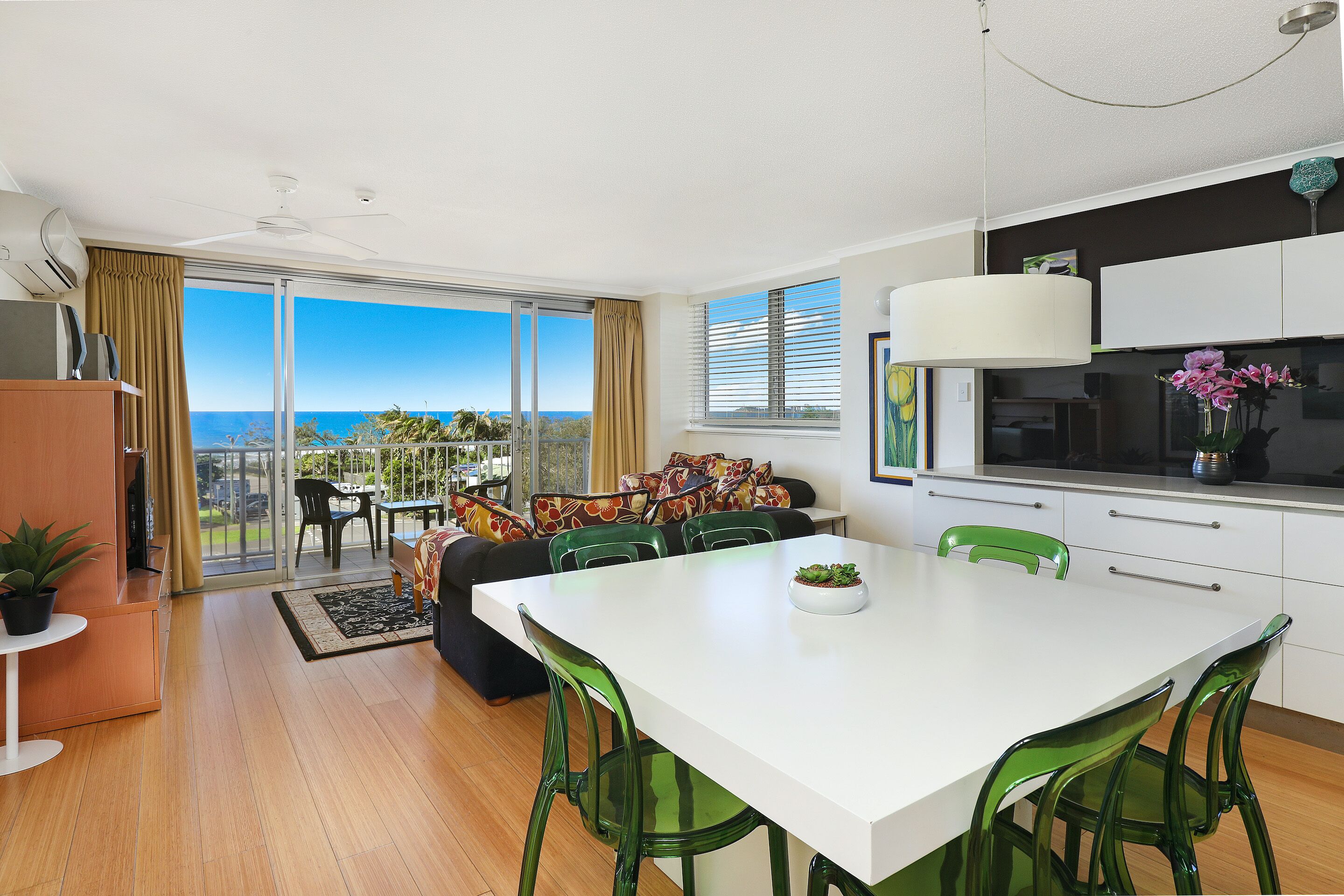 A modern kitchen with a white countertop and green chairs, a dining area with a white table, and a balcony with a view of the ocean.