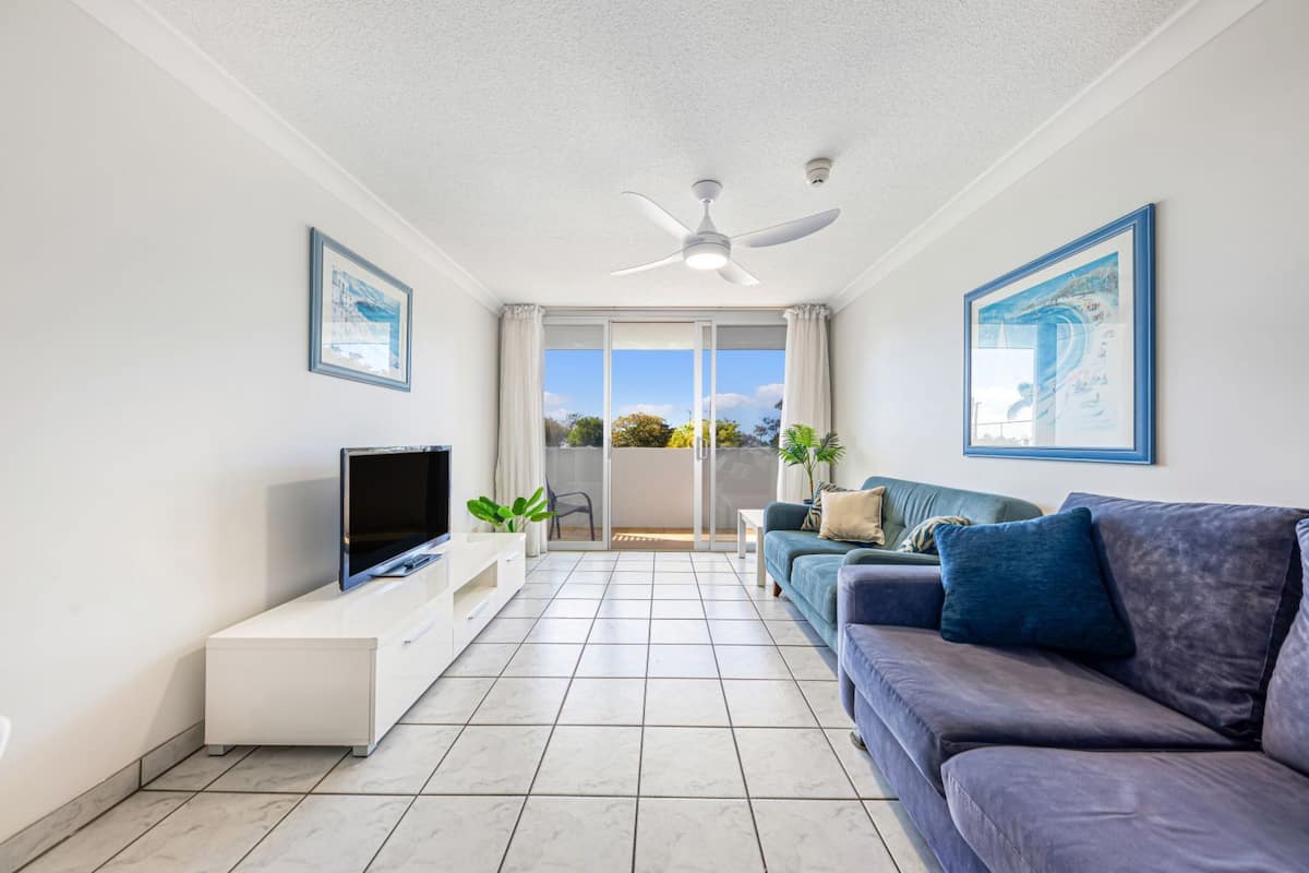 A modern living room with a flat-screen TV, a white entertainment unit, a blue sofa, and a ceiling fan.