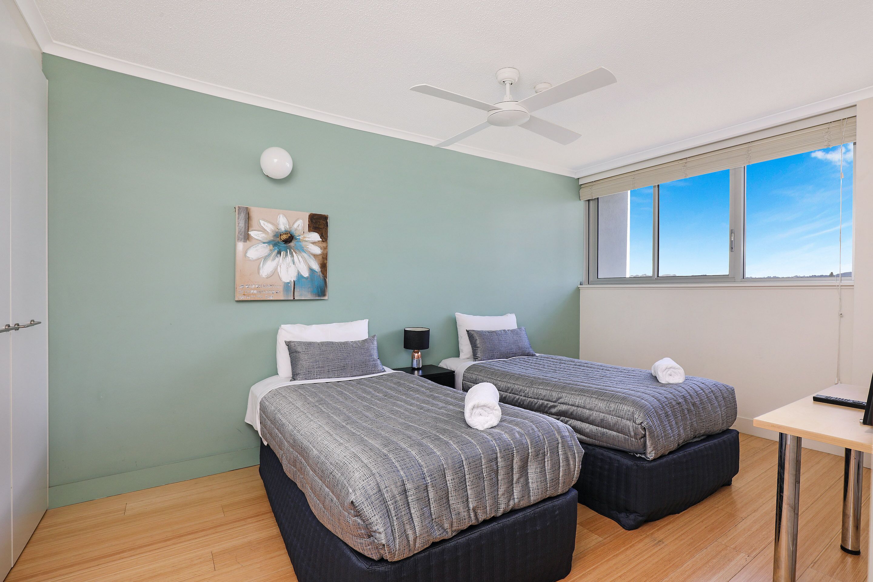A room with two beds, a ceiling fan, a painting, a desk, and a window with a view.