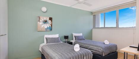 A room with two beds, a ceiling fan, a painting, a desk, and a window with a view.