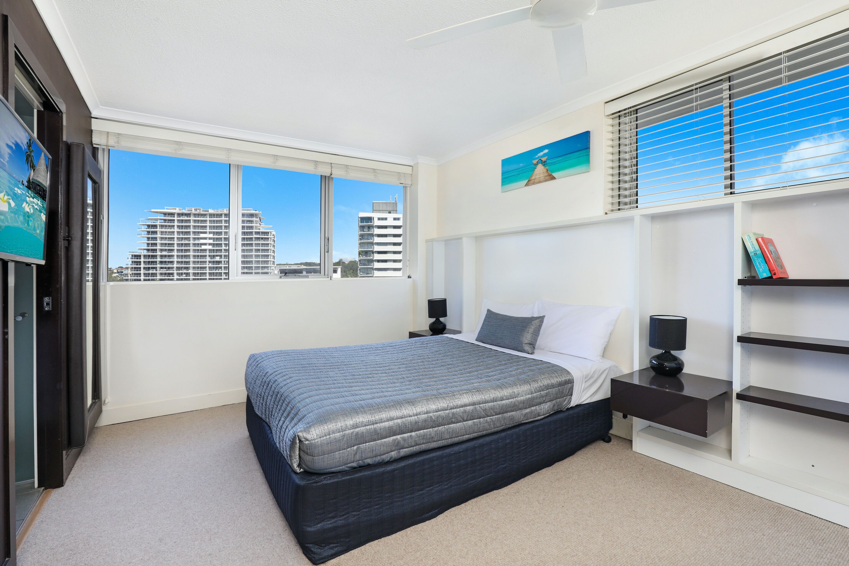 Un dormitorio con una cama grande, una mesita de noche, un estante en la pared y una ventana con vista a la ciudad.