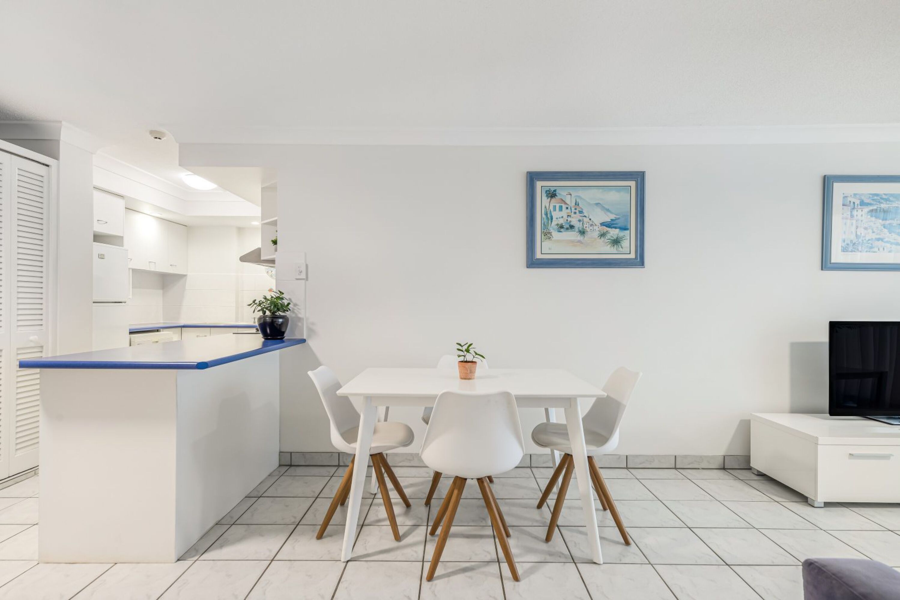 A modern kitchen with white cabinets and a blue countertop, a dining area with white chairs, and a TV on a white cabinet.