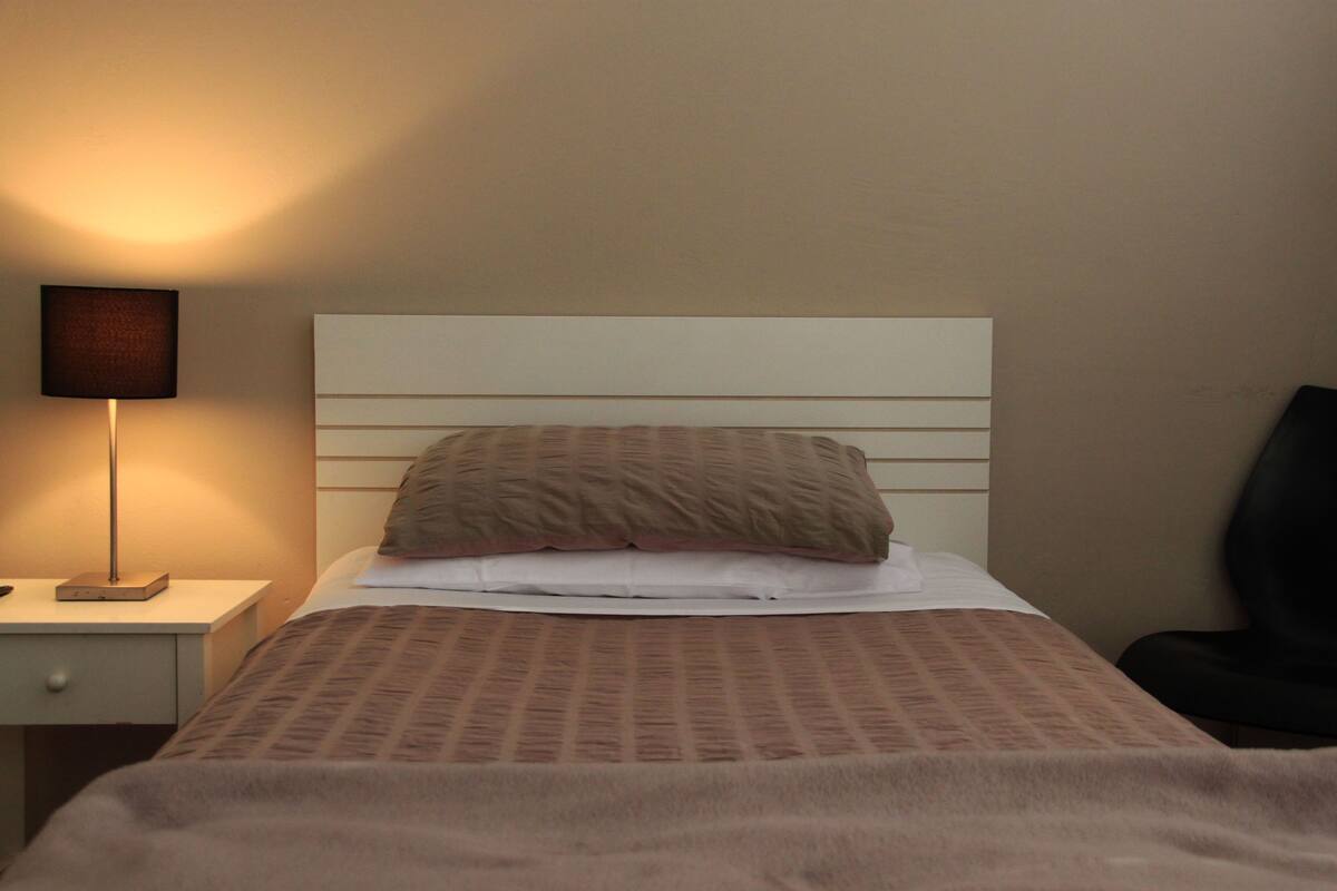 A neatly made bed with a patterned blanket, a bedside lamp, and a chair to the right.