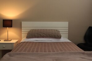 Una cama bien hecha con un edredón estampado, una lámpara de noche y una silla a la derecha.