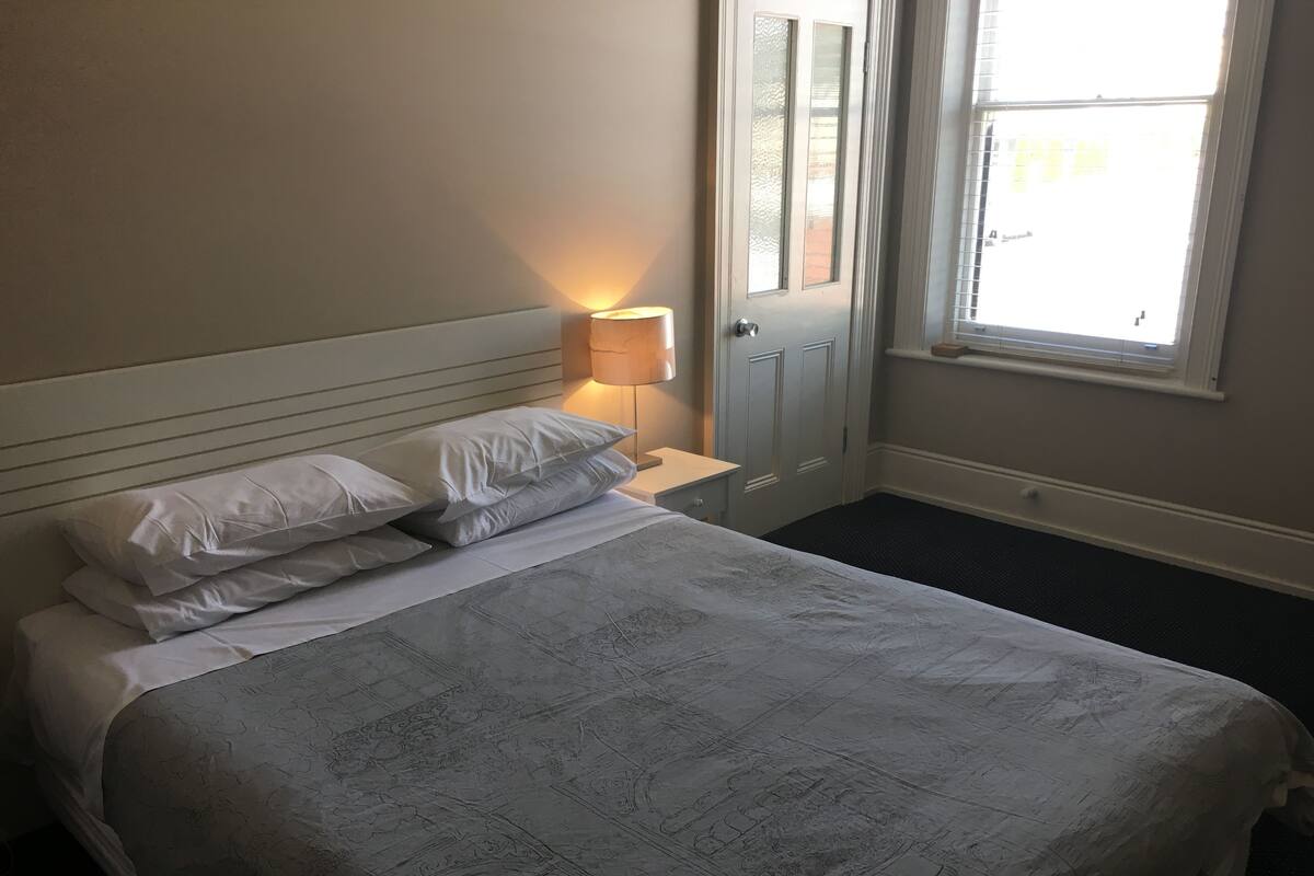 A neatly made bed with a patterned bedspread, a bedside lamp, and a window with blinds.