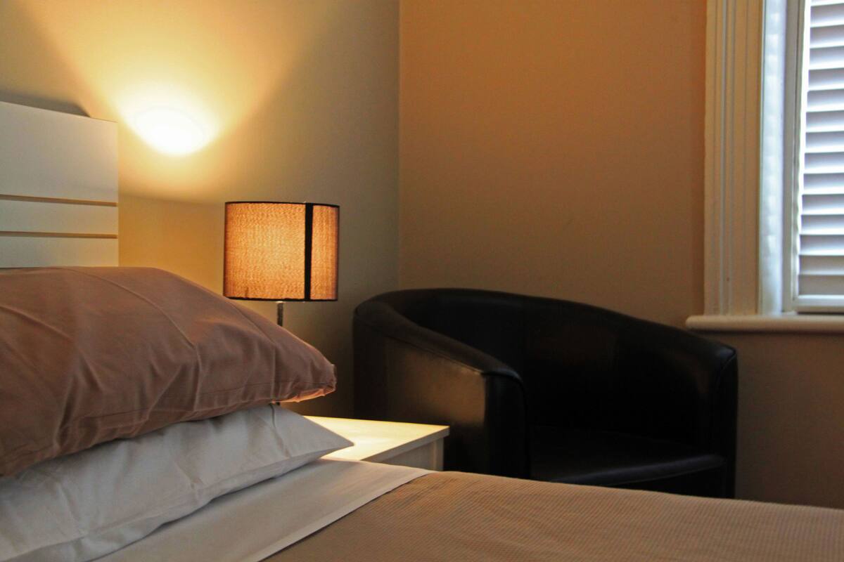 A hotel room with a bed, a chair, a lamp, and a window with blinds.