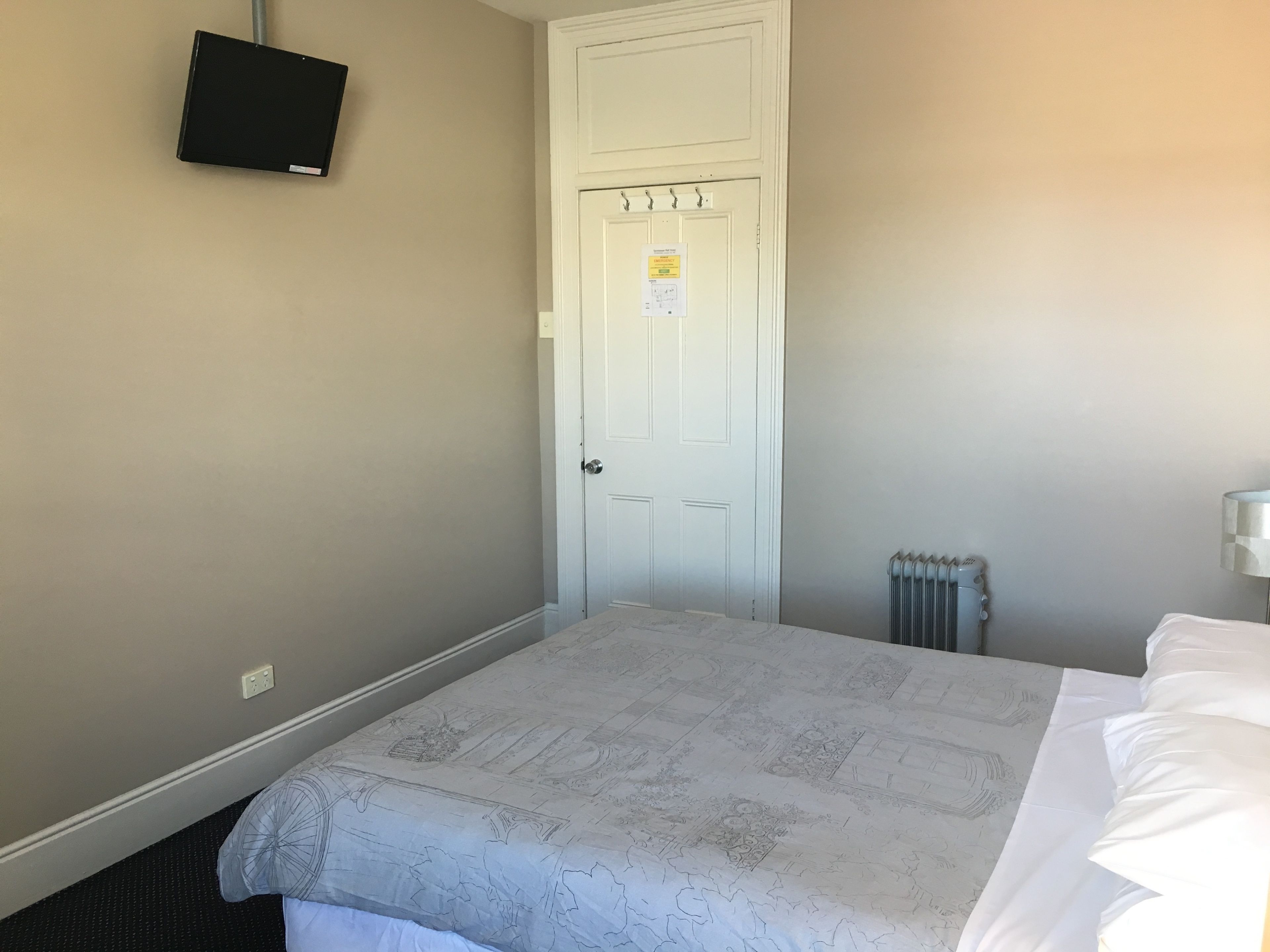 Une chambre d’hôtel avec un lit, une porte blanche, un radiateur et un téléviseur fixé au mur.