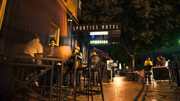 Un área de estar al aire libre en el Hotel Sporties con mesas y sillas, un letrero sobre la entrada y un árbol de fondo.