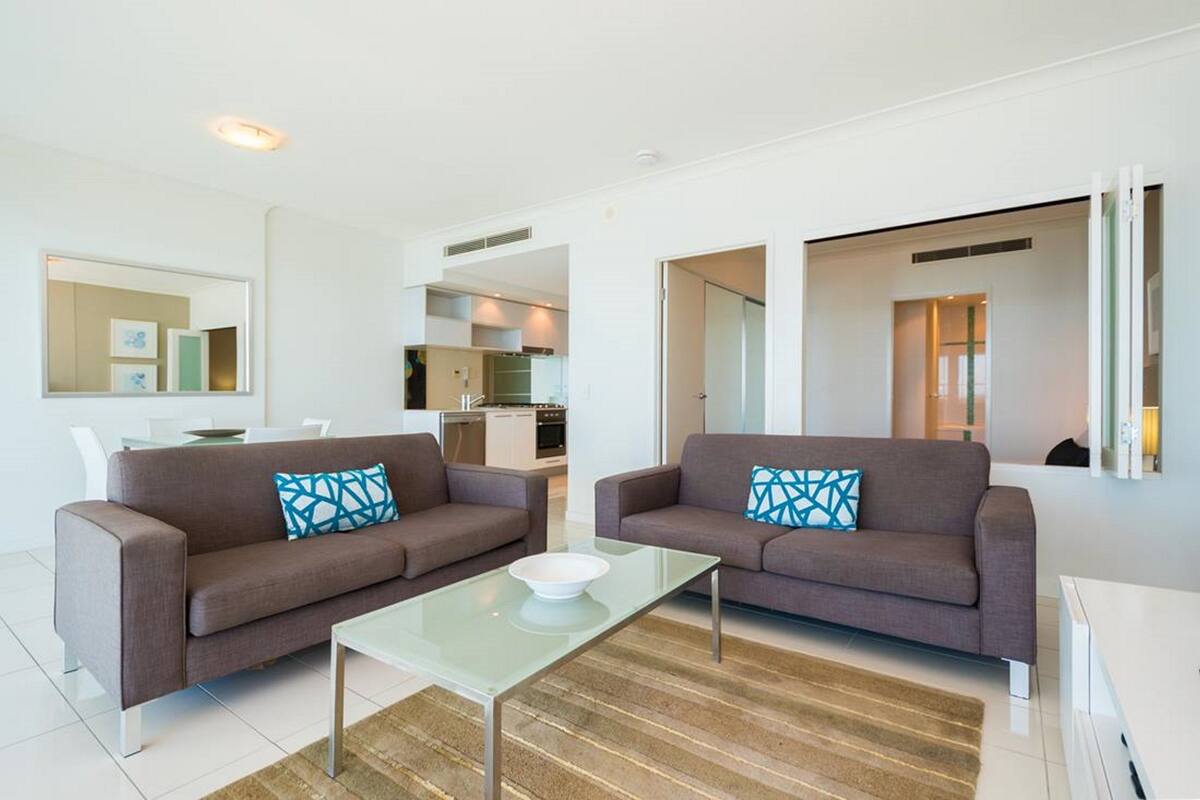 Ein modernes Wohnzimmer mit zwei Sofas, einem Glastisch und einer Küche im Hintergrund.