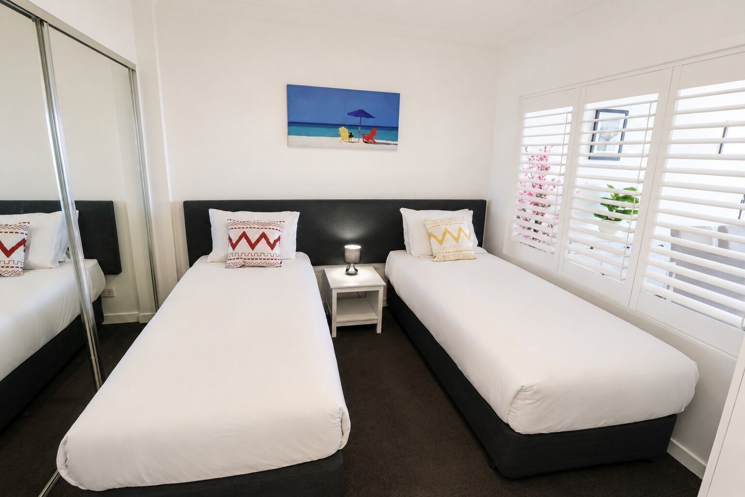 A hotel room with two single beds, a nightstand, a lamp, a painting of a beach scene, and a window with blinds.