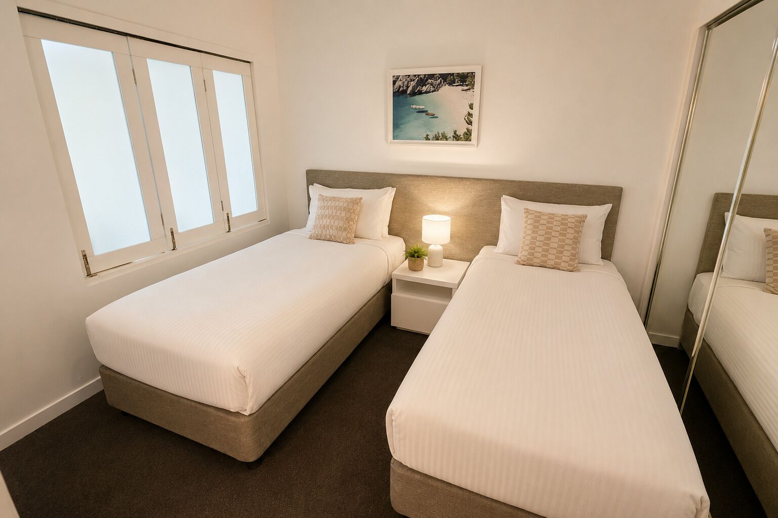 Quarto de hotel com duas camas de solteiro, mesa de cabeceira com abajur, uma pequena planta em vaso e um quadro na parede.