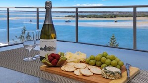 Une bouteille de vin, des verres, du fromage, des raisins et des crackers sont posés sur une table, avec une vue sur une plage et l’océan.