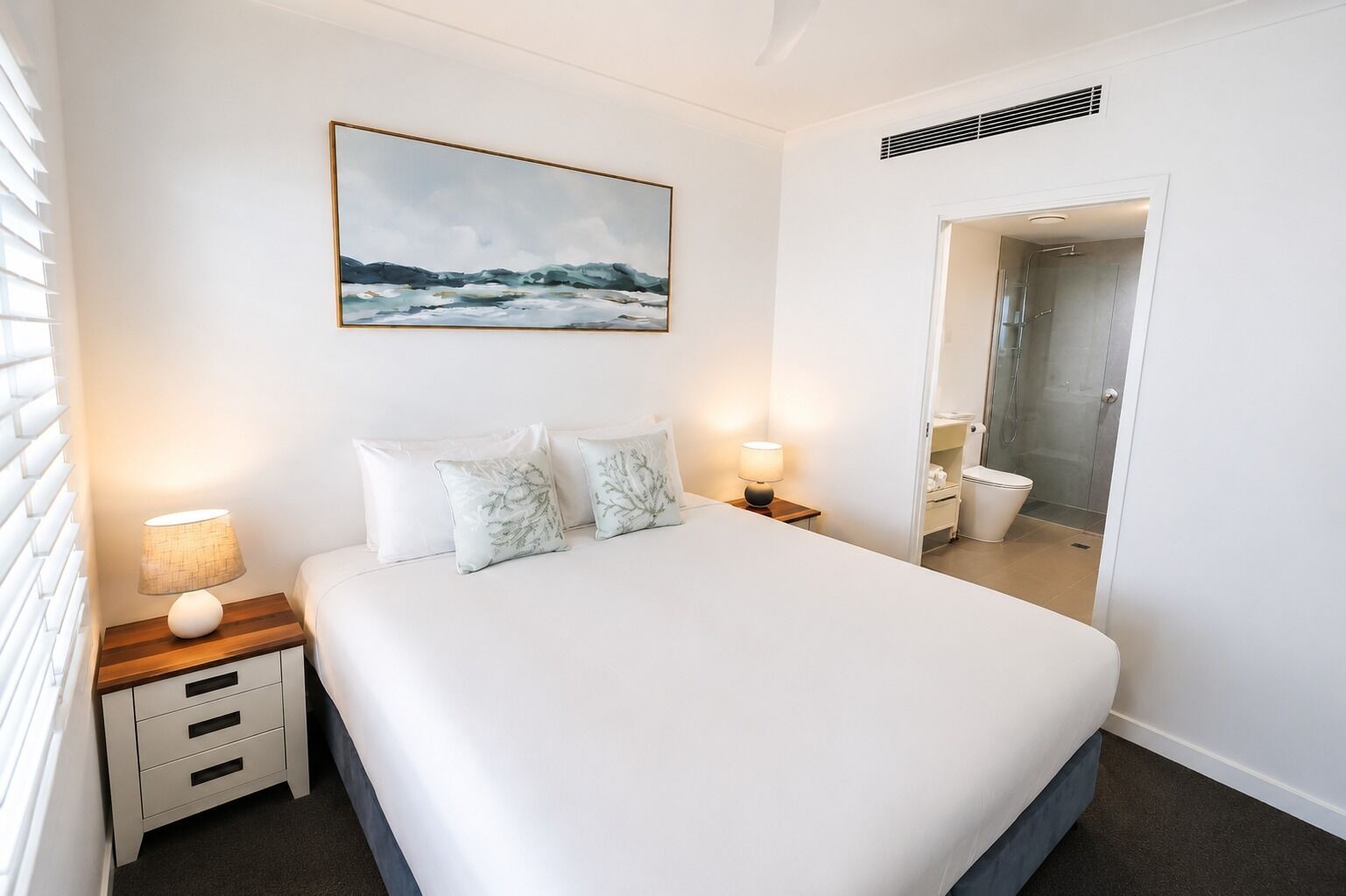 A hotel room with a large bed, bedside lamps, a painting, and a bathroom visible through an open door.