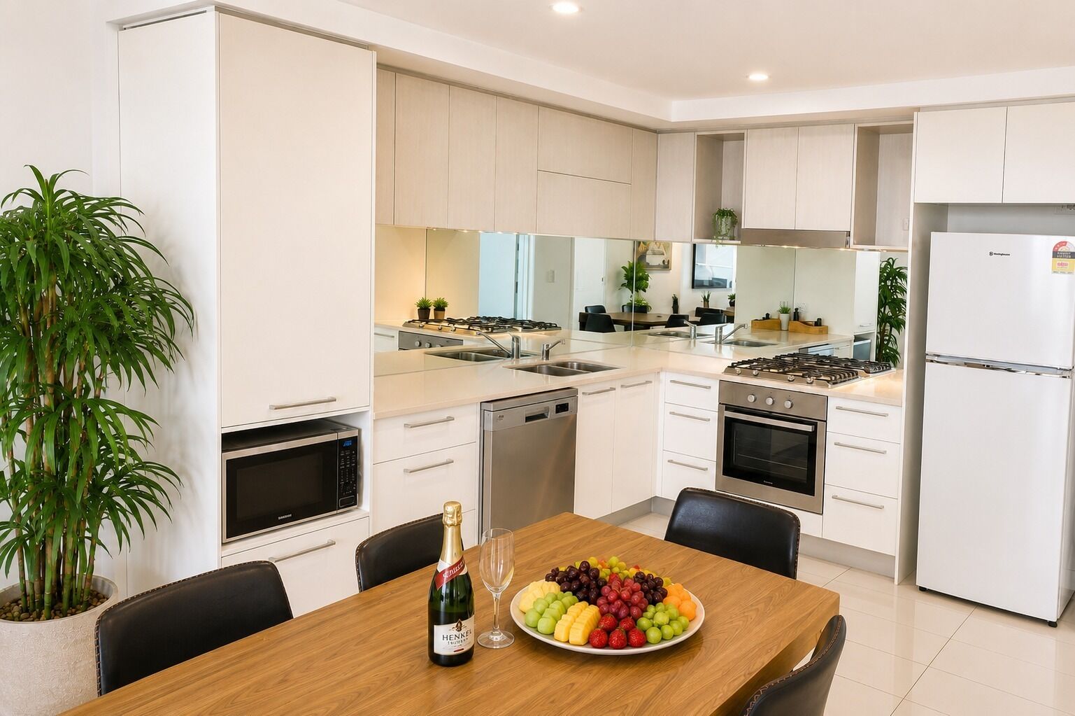 A modern kitchen with white cabinetry, a dining table set with chairs, a bottle of wine, and a fruit platter.