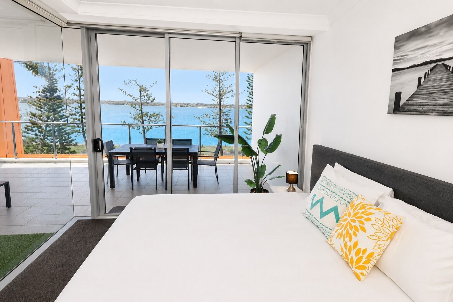 Un dormitorio con una cama grande, vista al mar a través de puertas de cristal, una zona de comedor con sillas y una mesa, y una planta en maceta.
