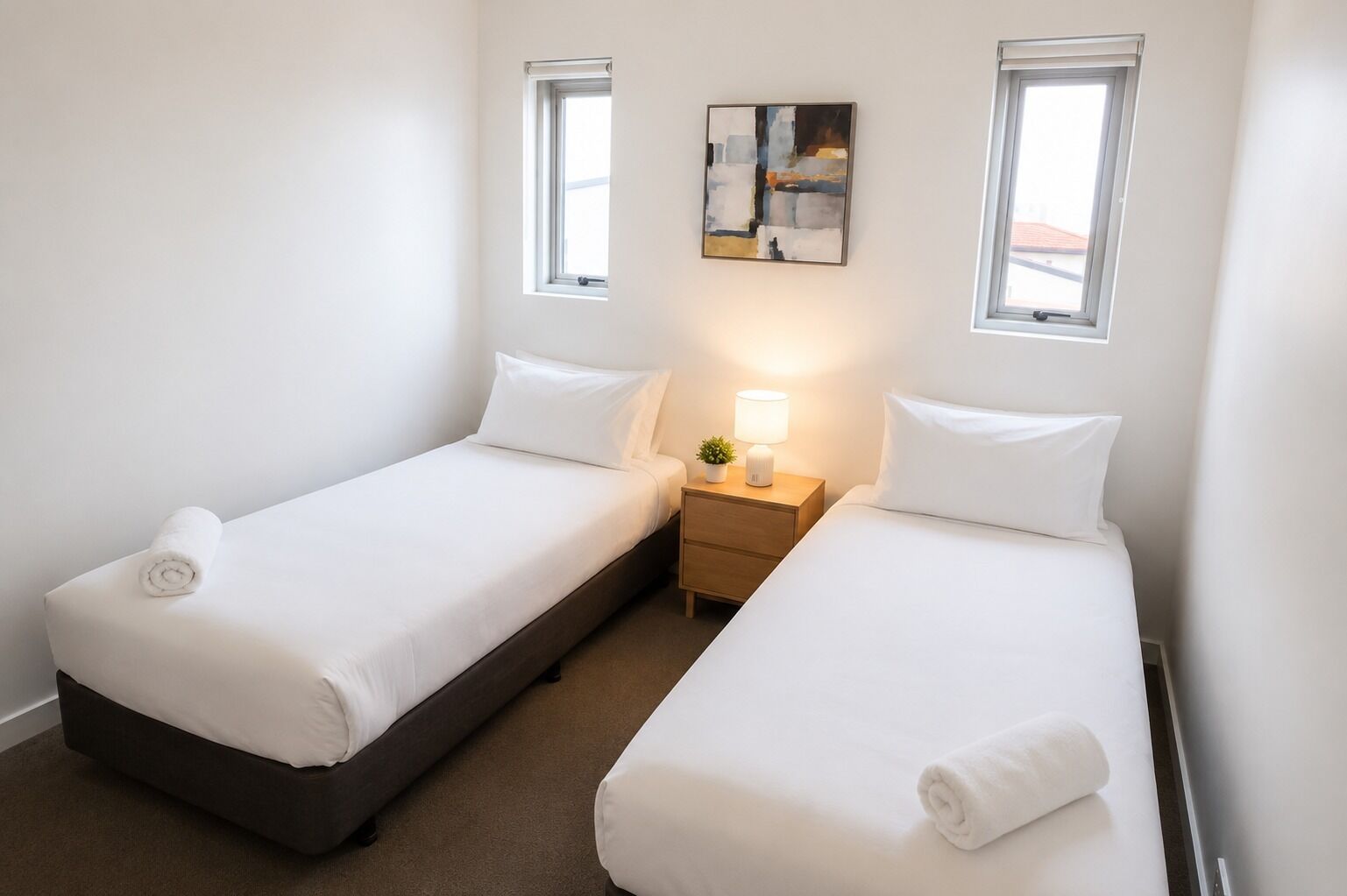 A small, clean room with two single beds, a nightstand with a lamp, and a painting on the wall.