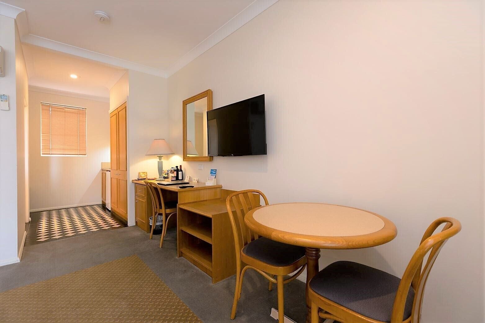 A hotel room with a desk, two chairs, a round table, a TV mounted on the wall, and a window with blinds.