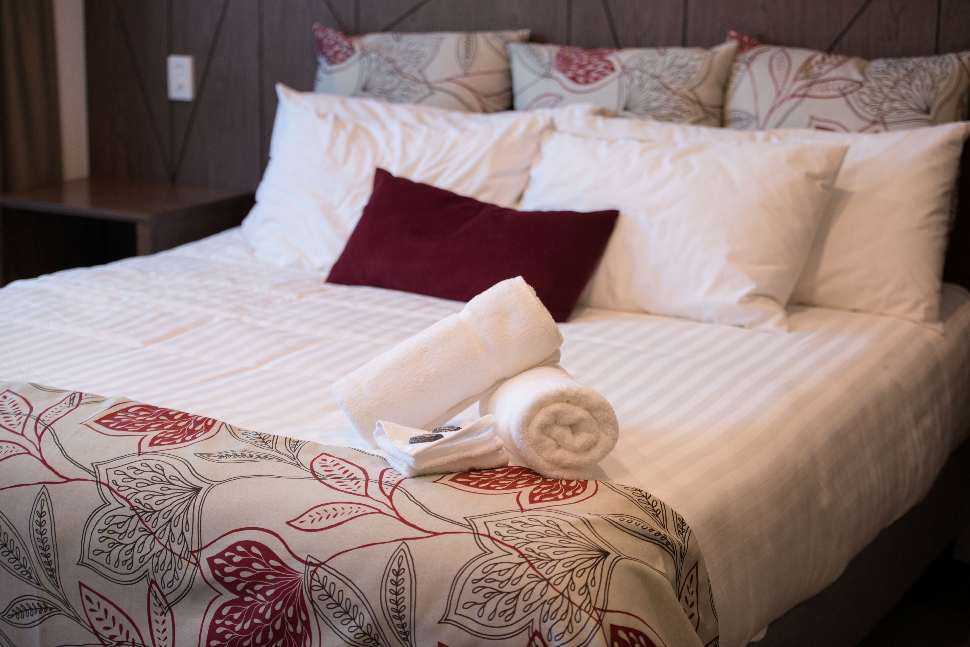 Una cama bien hecha con sábanas blancas, una almohada roja y toallas enrolladas.