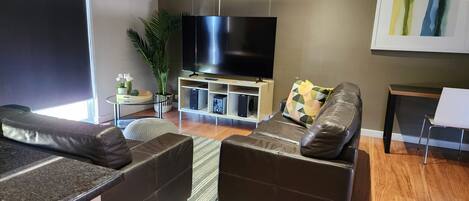 A modern living room with a large flat-screen TV, leather sofas, a small side table with a plant, and a framed abstract painting on the wall.