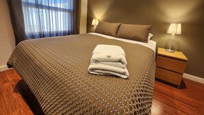 Cama bem arrumada com colcha estampada, toalha dobrada, duas luminárias de cabeceira e uma janela com cortinas finas.