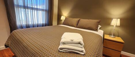 A neatly made bed with a patterned bedspread, a folded towel, two bedside lamps, and a window with sheer curtains.