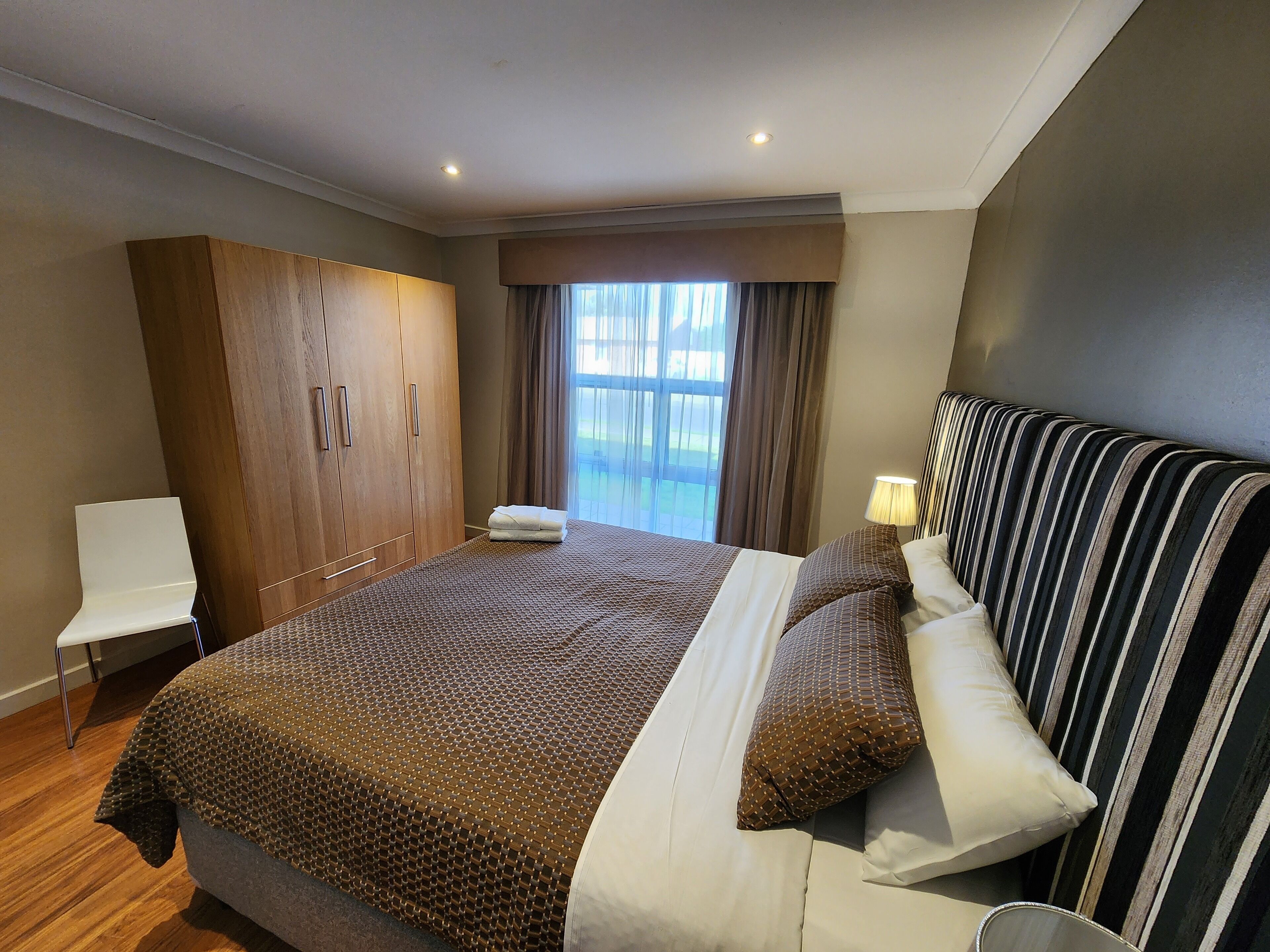 A modern hotel room with a large bed, a wooden wardrobe, a white chair, and a view of the outdoors through the window.