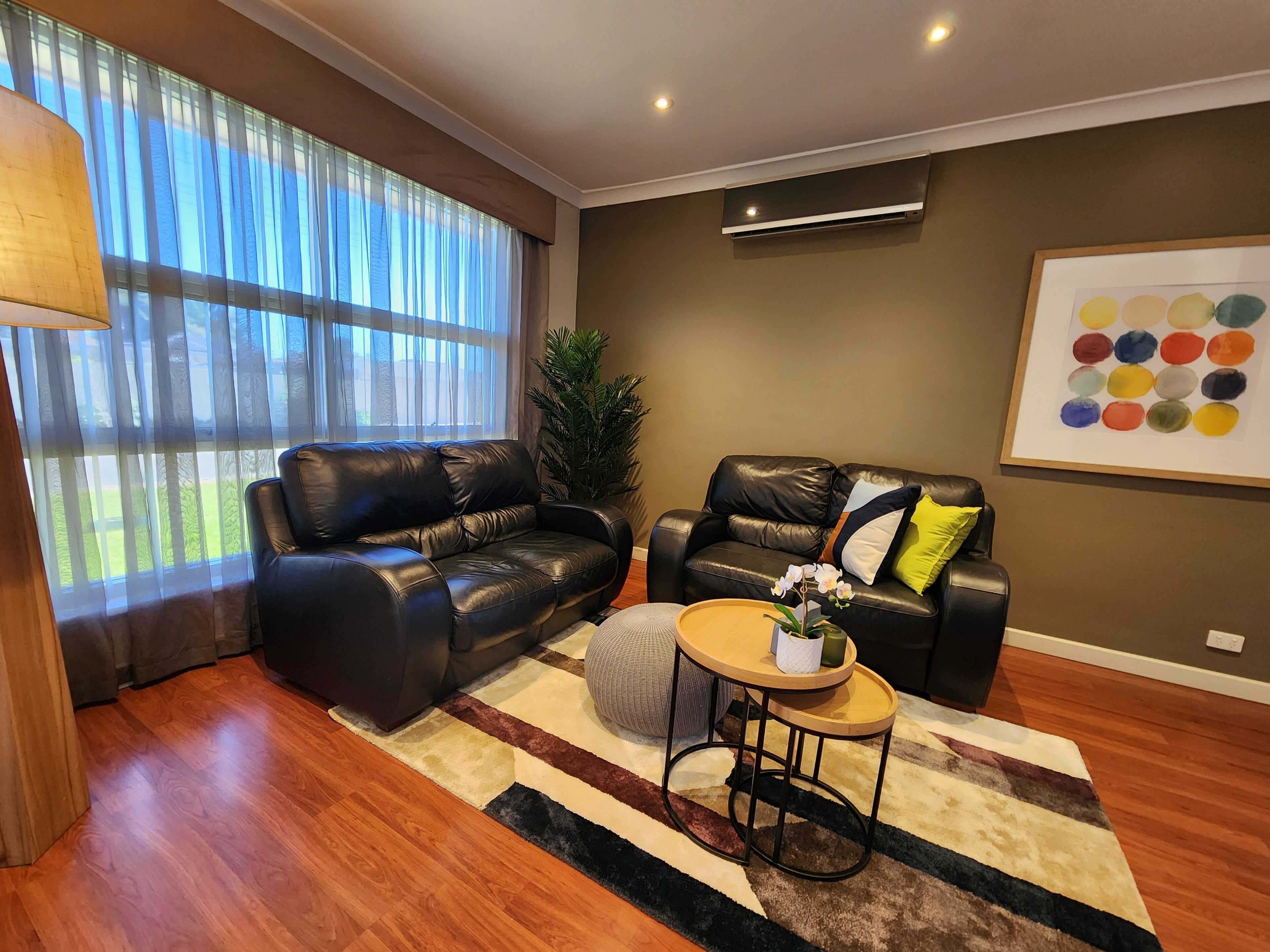 A modern living room with a black leather sofa, a round wooden coffee table, and a large window with sheer curtains.