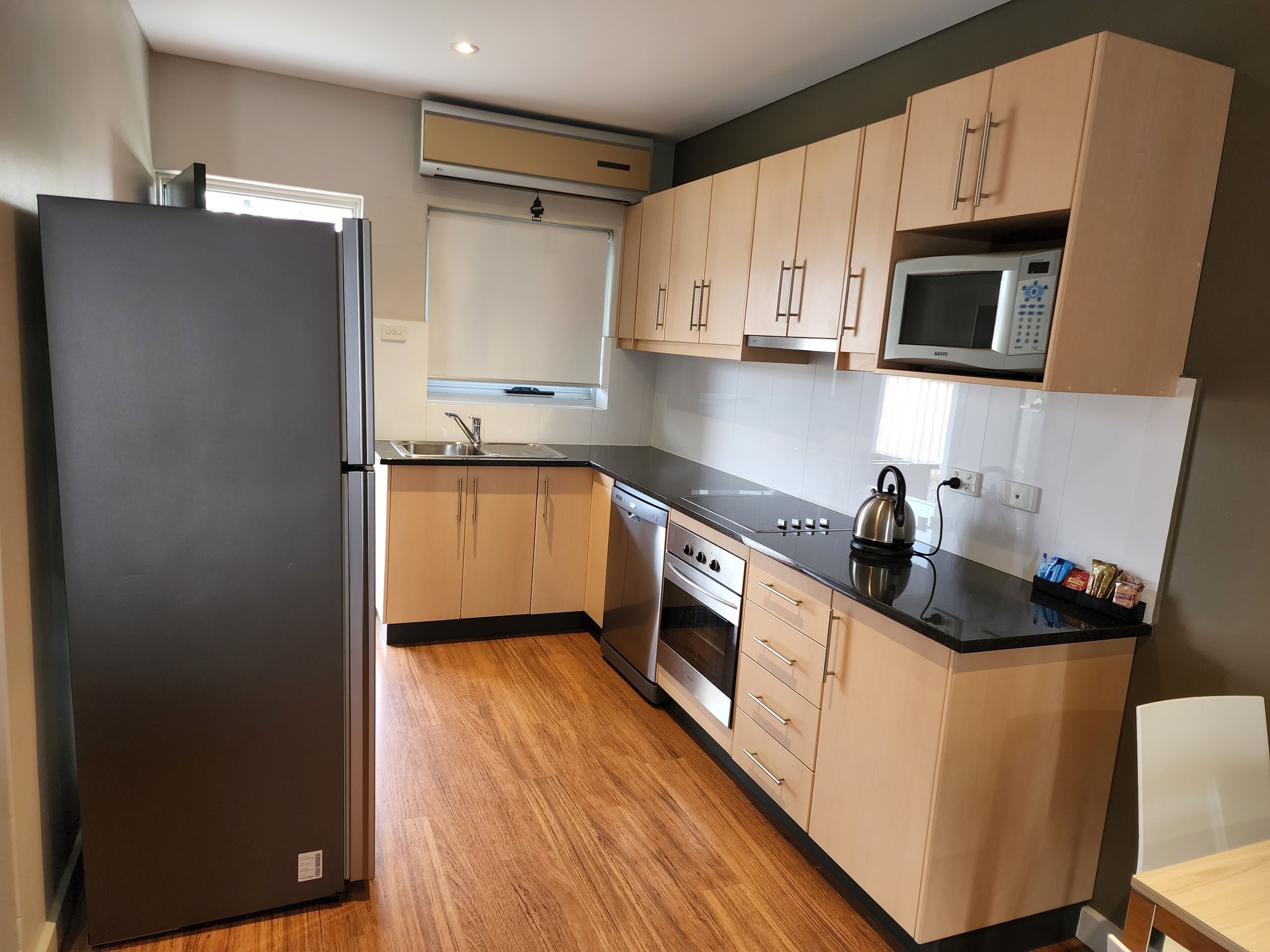 A modern kitchen with wooden cabinets, a stainless steel refrigerator, an oven, a microwave, and a kettle.