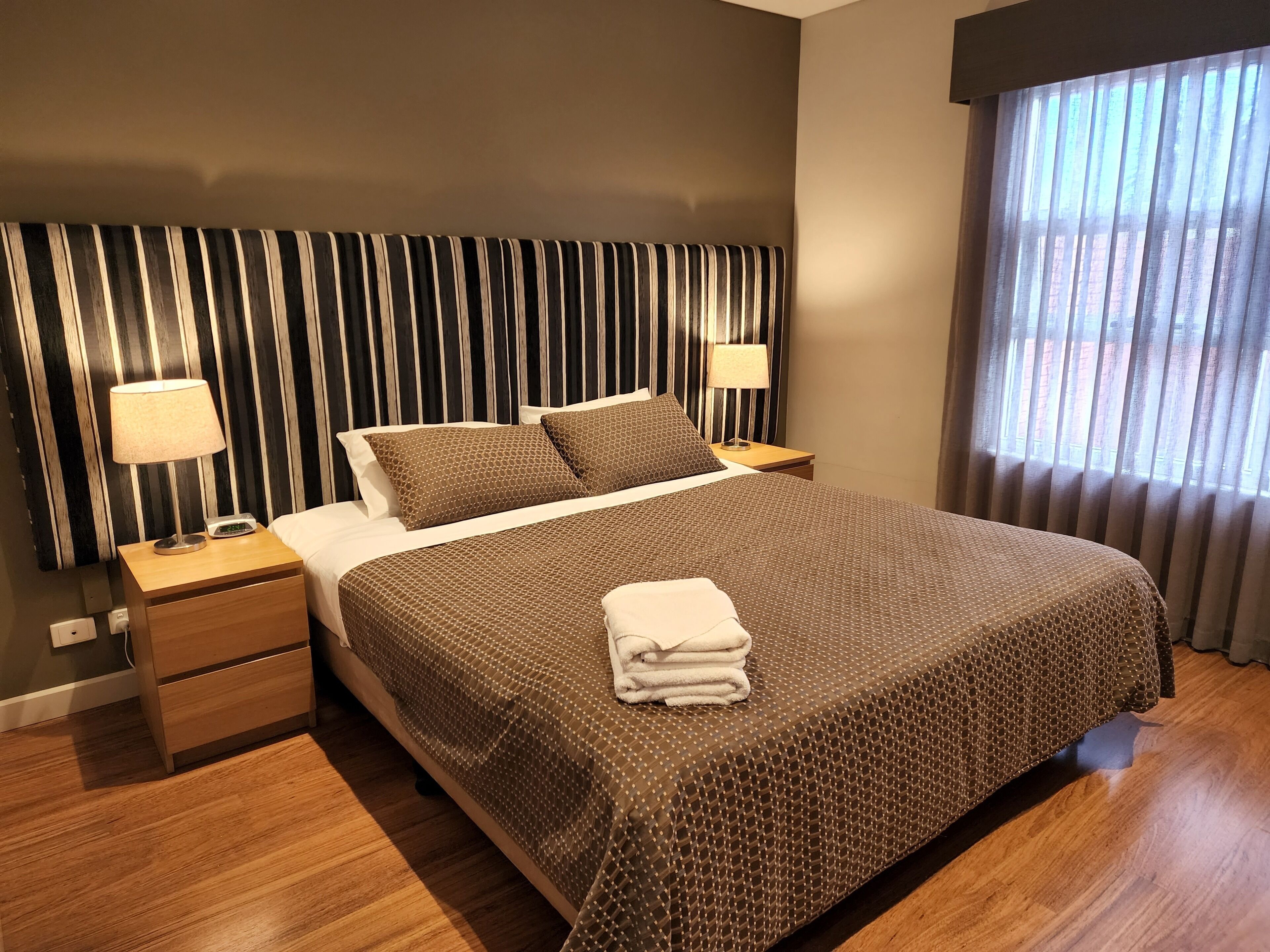 A hotel room with a large bed, two bedside tables with lamps, a wooden floor, and a window with curtains.