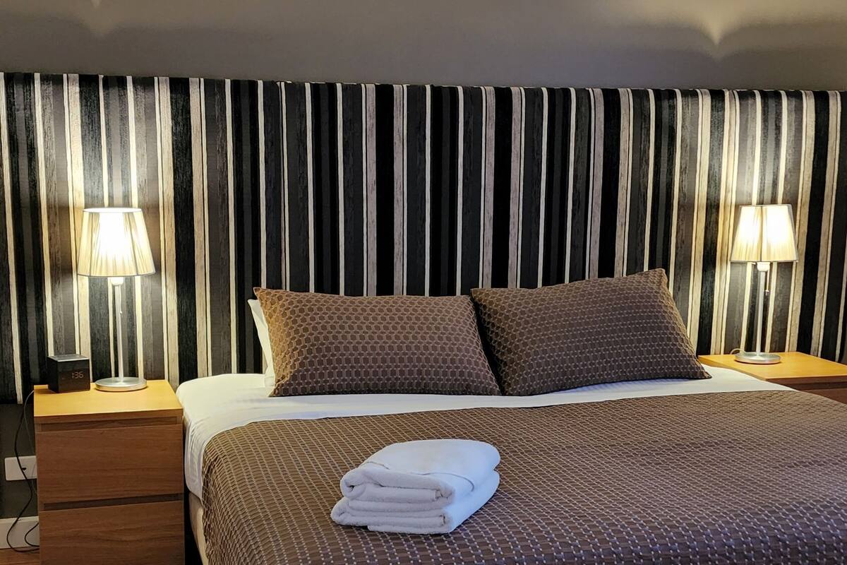 A neatly made bed with a patterned bedspread and two pillows, flanked by two bedside tables with lamps, against a striped curtain backdrop.