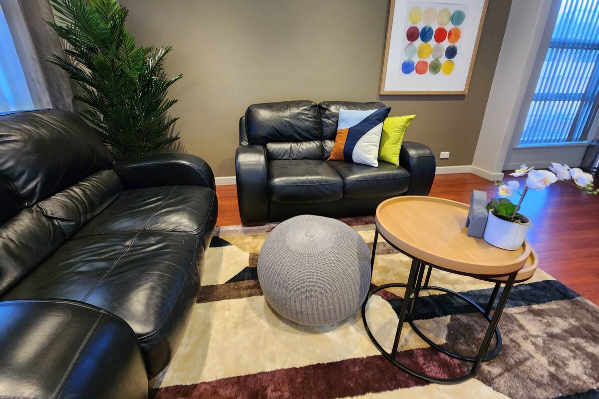 A living room with a black leather sofa, a round ottoman, and a coffee table. There is a potted plant on the coffee table.
