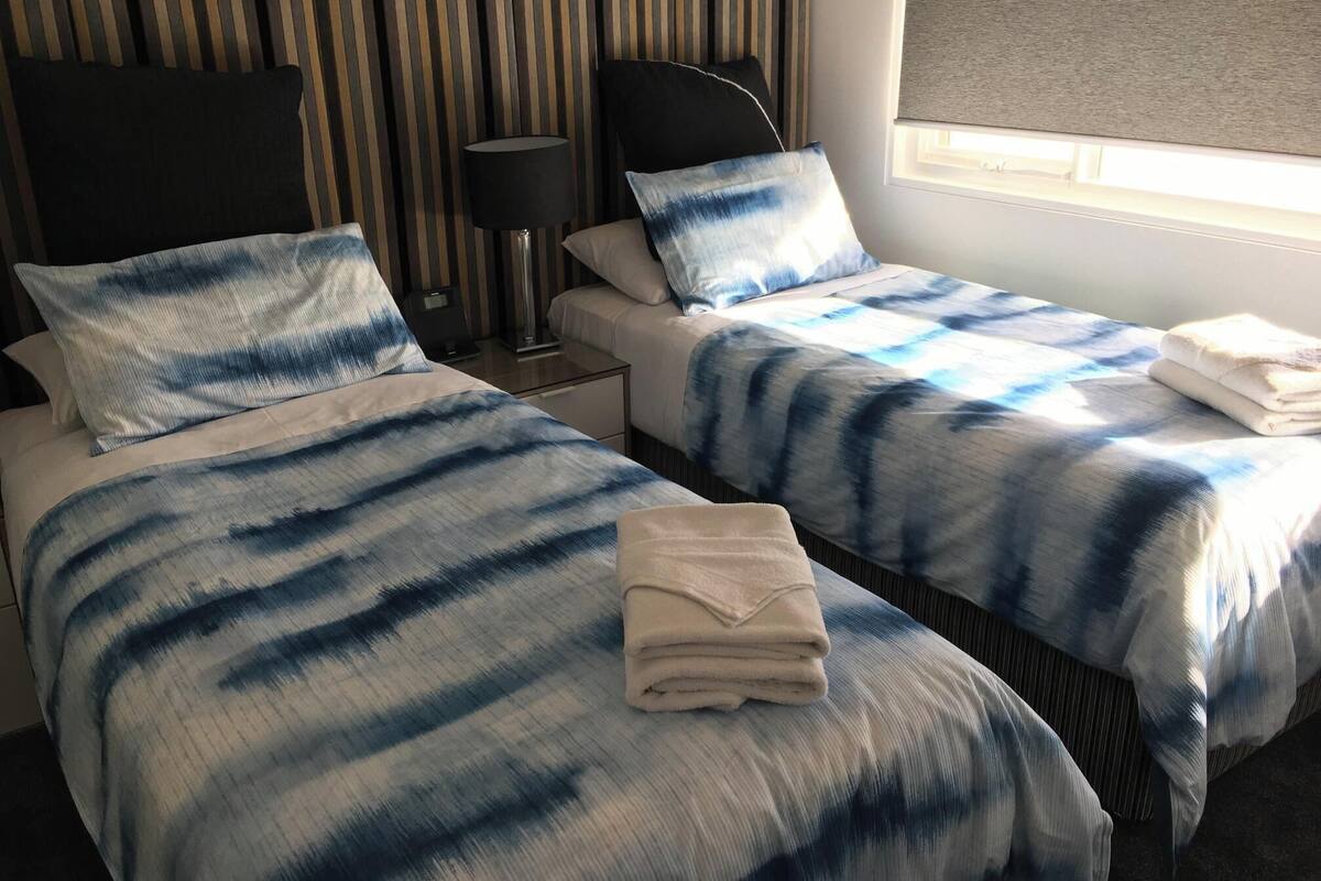 A hotel room with two beds, striped headboards, a bedside table with a lamp, and a window with a blind.