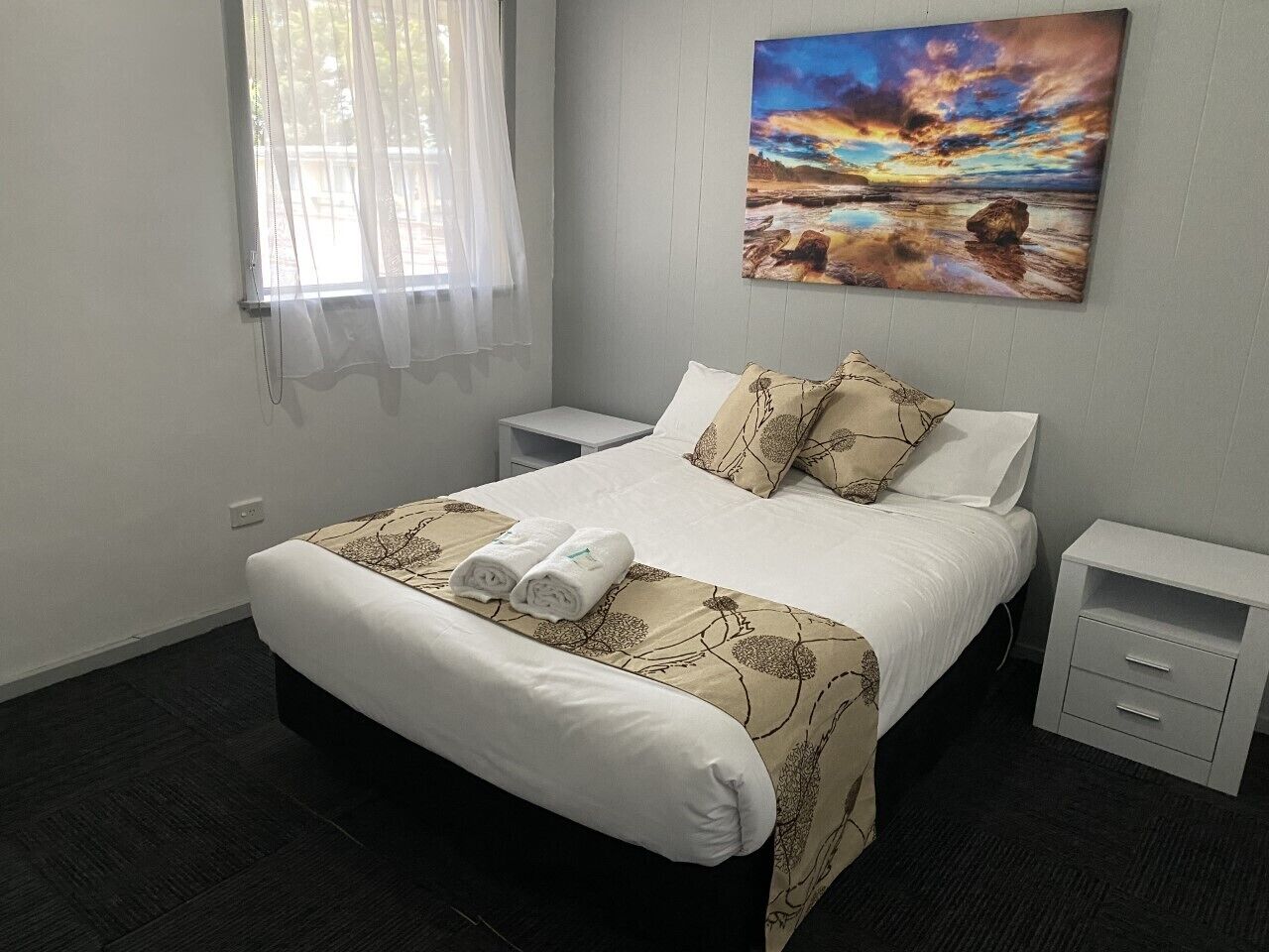 Cama bem arrumada com colcha floral, dois travesseiros e duas toalhas enroladas. Tem uma mesa de cabeceira com gaveta e um quadro com um pôr do sol na parede.