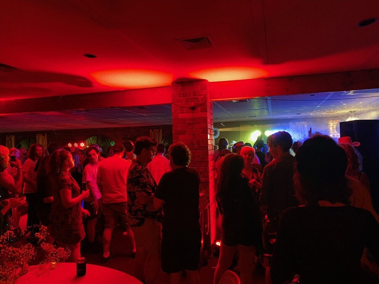 A crowded indoor event with people mingling, a brick pillar, and a stage with lighting in the background.