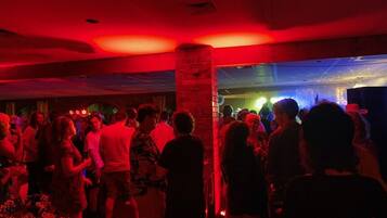 A crowded indoor event with people mingling, a brick pillar, and a stage with lighting in the background.