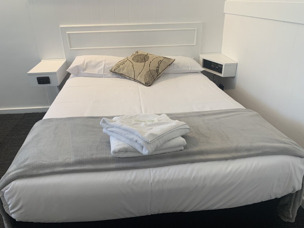 A neatly made bed with white linens, a grey blanket, and a decorative pillow. There is a small bedside table with a lamp and a power outlet.
