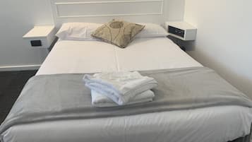 A neatly made bed with white linens, a grey blanket, and a decorative pillow. There is a small bedside table with a lamp and a power outlet.