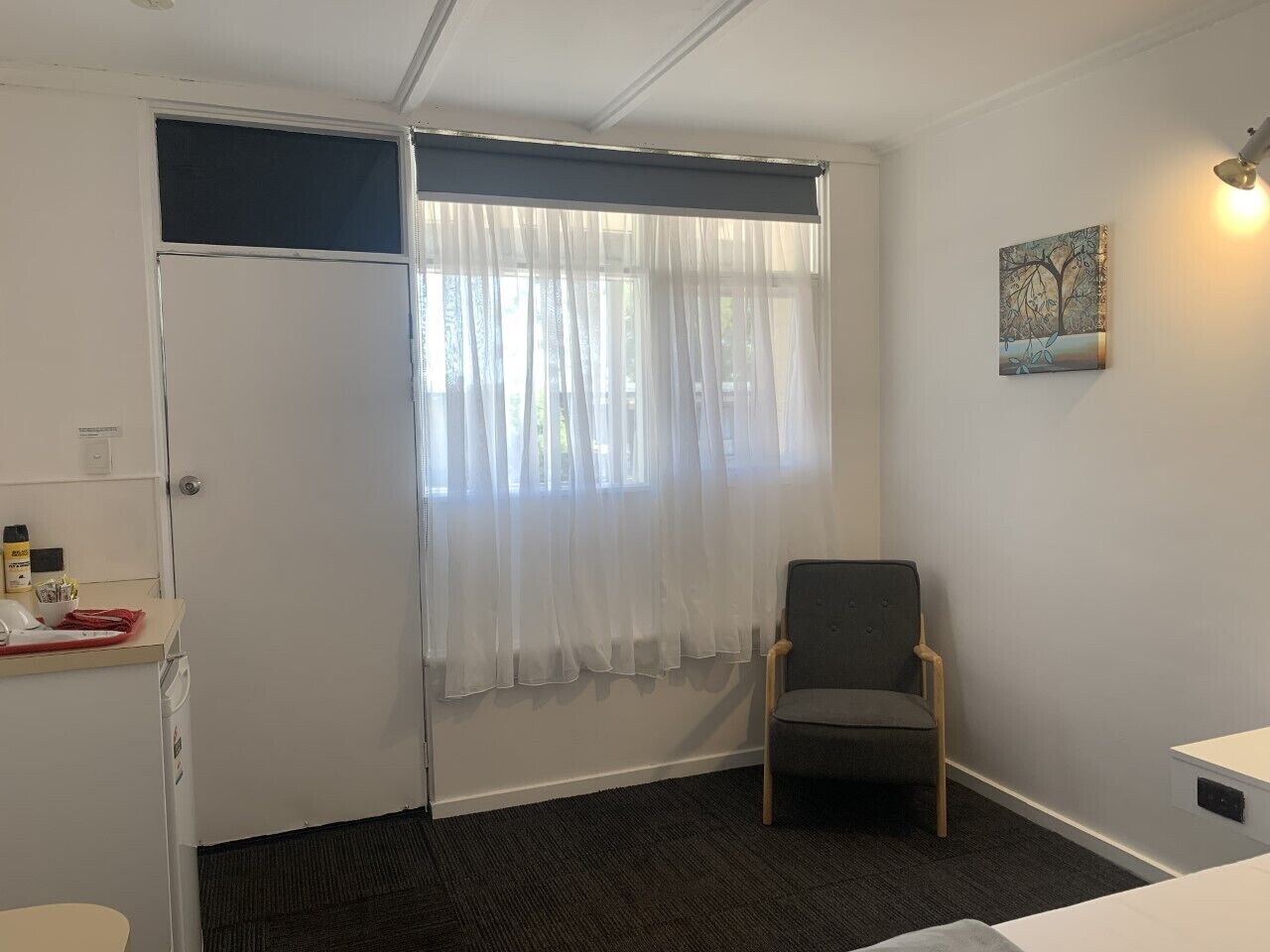 Ein Zimmer mit einem Bett, einem Sessel, einem Fenster mit leichtem Vorhang, einem Bild an der Wand und einem kleinen Tisch mit Gegenständen darauf.