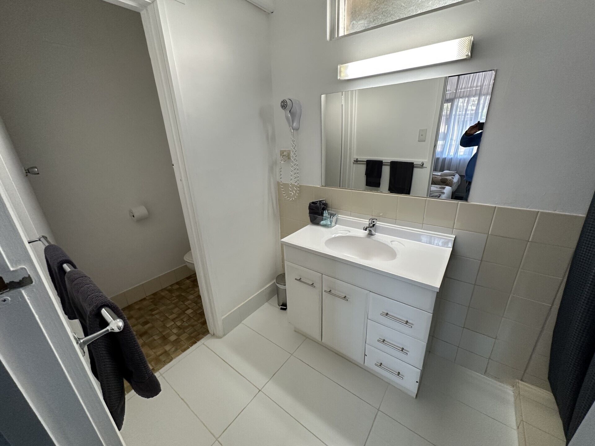 Un baño con lavamanos blanco, espejo y paredes con azulejos. Se ve un perchero con una toalla, y una zona de ducha con cortina.
