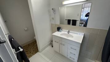 Un baño con lavamanos blanco, espejo y paredes con azulejos. Se ve un perchero con una toalla, y una zona de ducha con cortina.