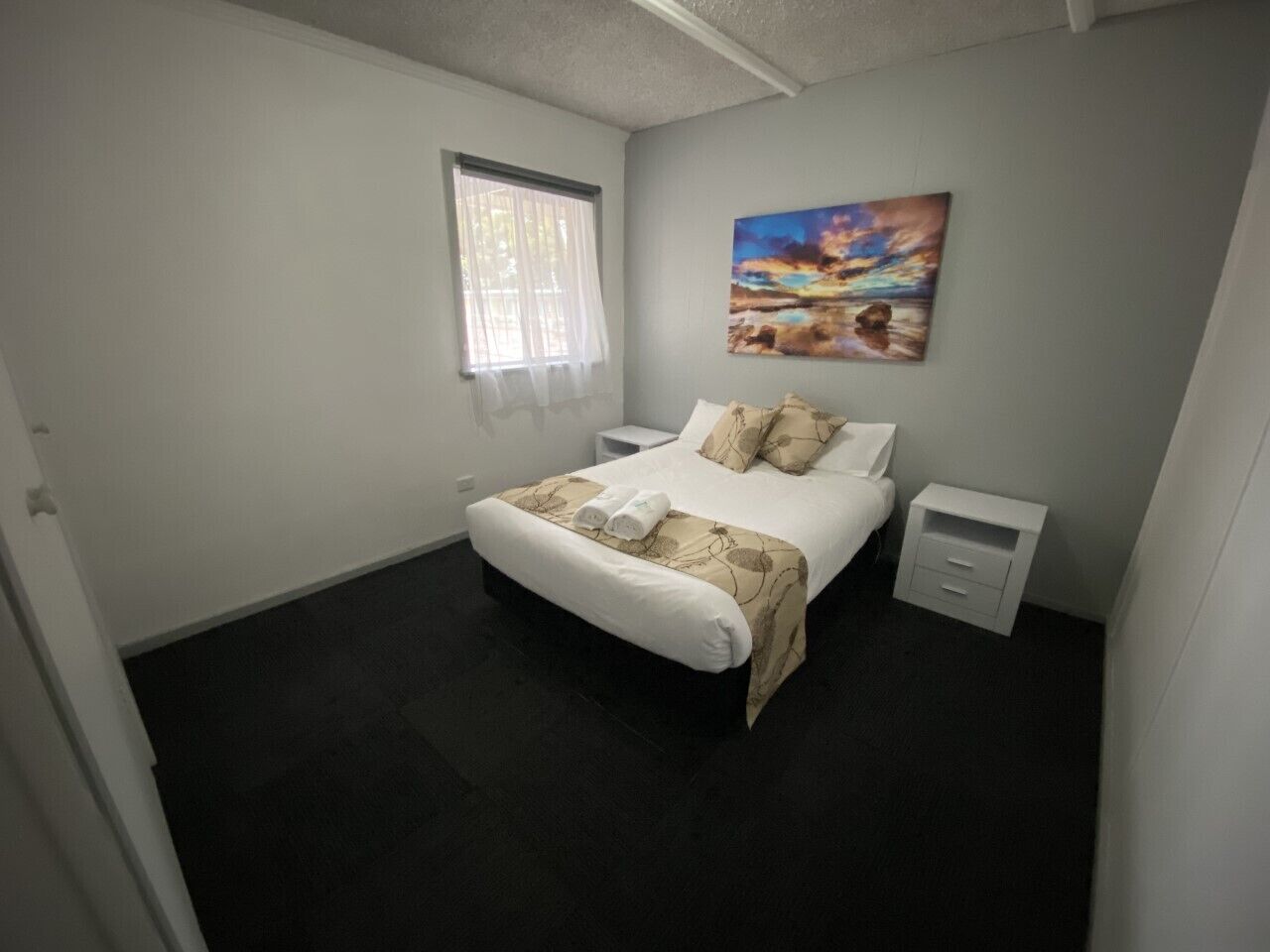 A bedroom with a bed, a nightstand, a window with curtains, and a painting on the wall.