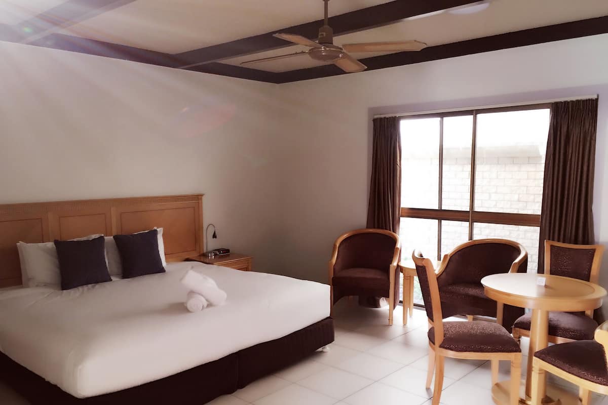 Habitación de hotel con una cama, dos sillas, una mesita, una mesita de noche y un ventanal con cortinas.