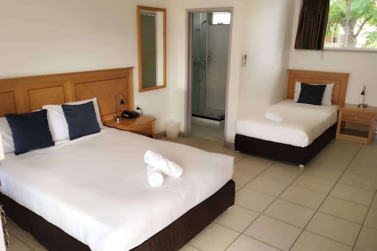 Una habitación de hotel con dos camas, un baño con puerta de vidrio y una ventana con cortinas.