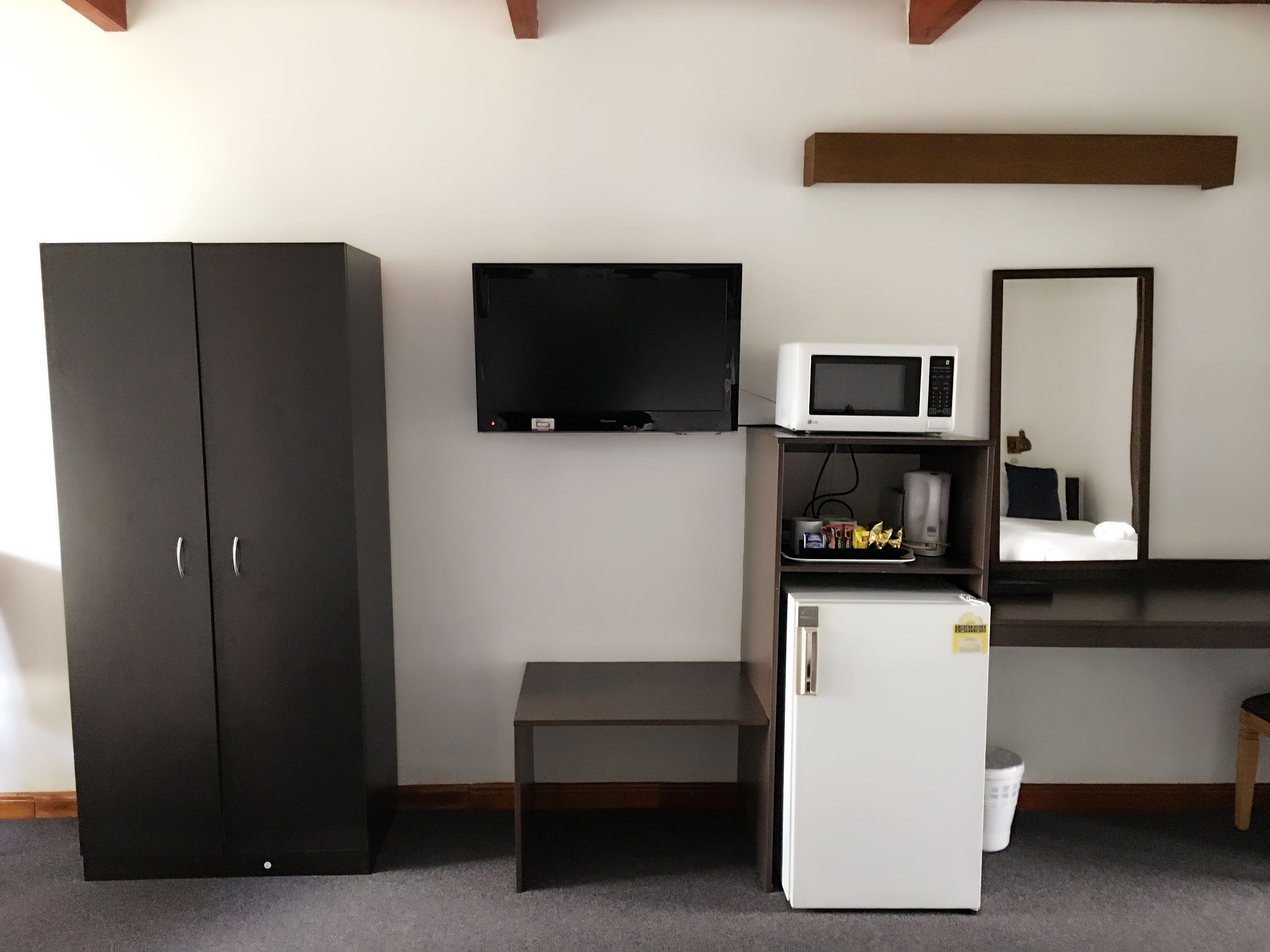 A compact room with a dark wardrobe, a flat-screen TV, a microwave, a small fridge, a bench, and a mirror.