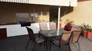 Un patio al aire libre con una mesa y sillas de vidrio, una parrilla empotrada y plantas en macetas.