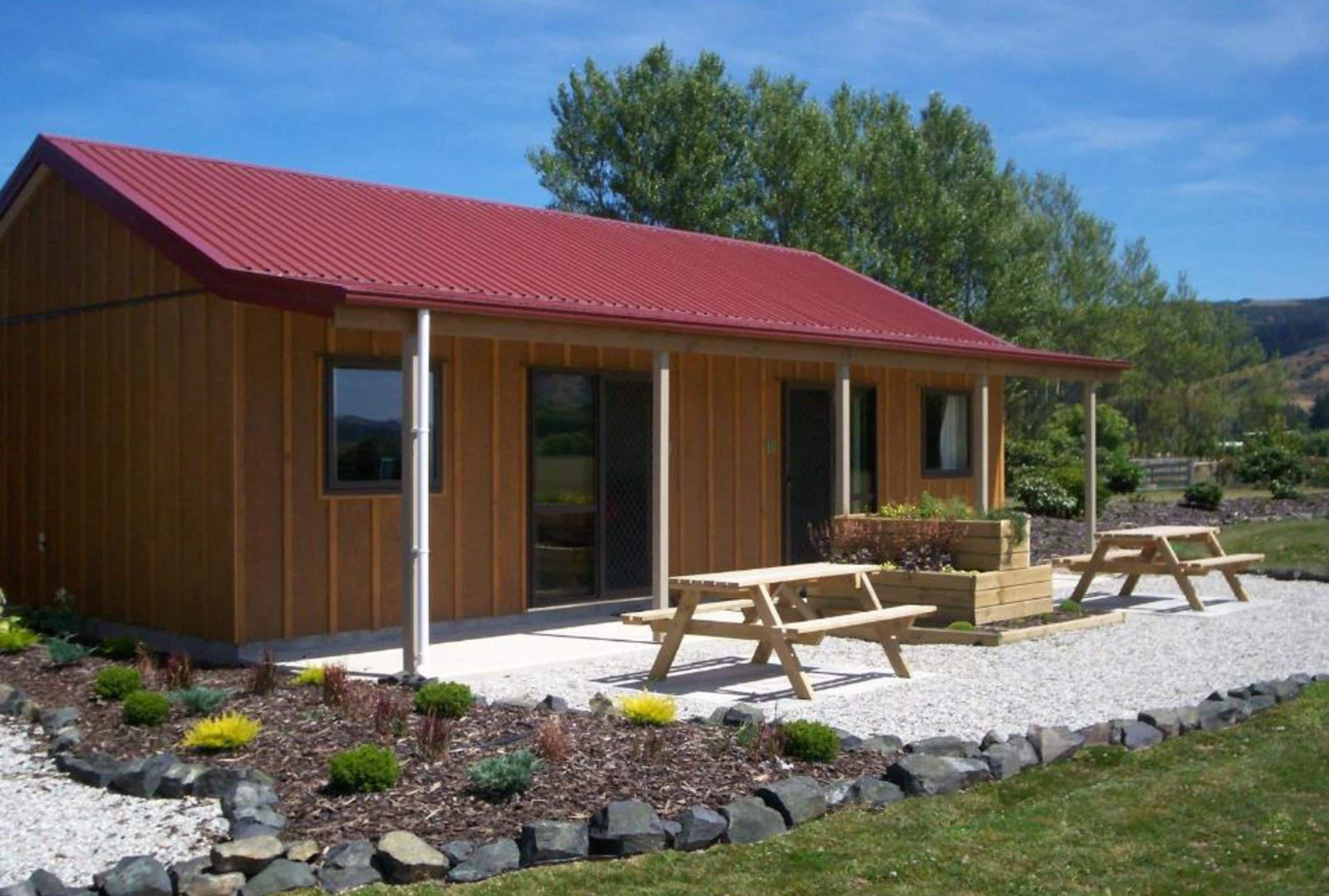 A wooden cabin with a red roof, a covered patio with picnic tables, and a gravel driveway.