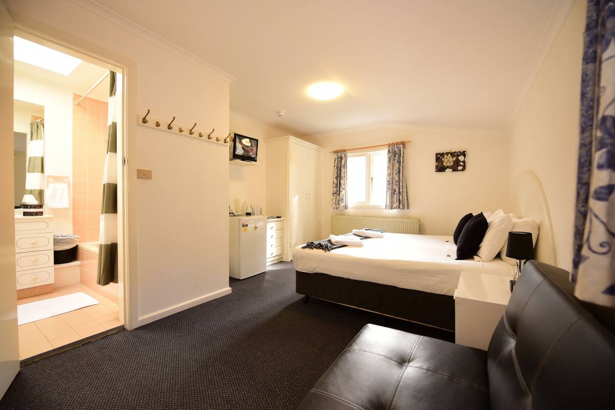 Quarto de hotel com cama, escrivaninha, cadeira, armário, banheiro e janela com cortinas.