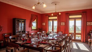 Une salle à manger avec une longue table dressée pour un repas, entourée de chaises, avec un buffet et des fenêtres en arrière-plan.