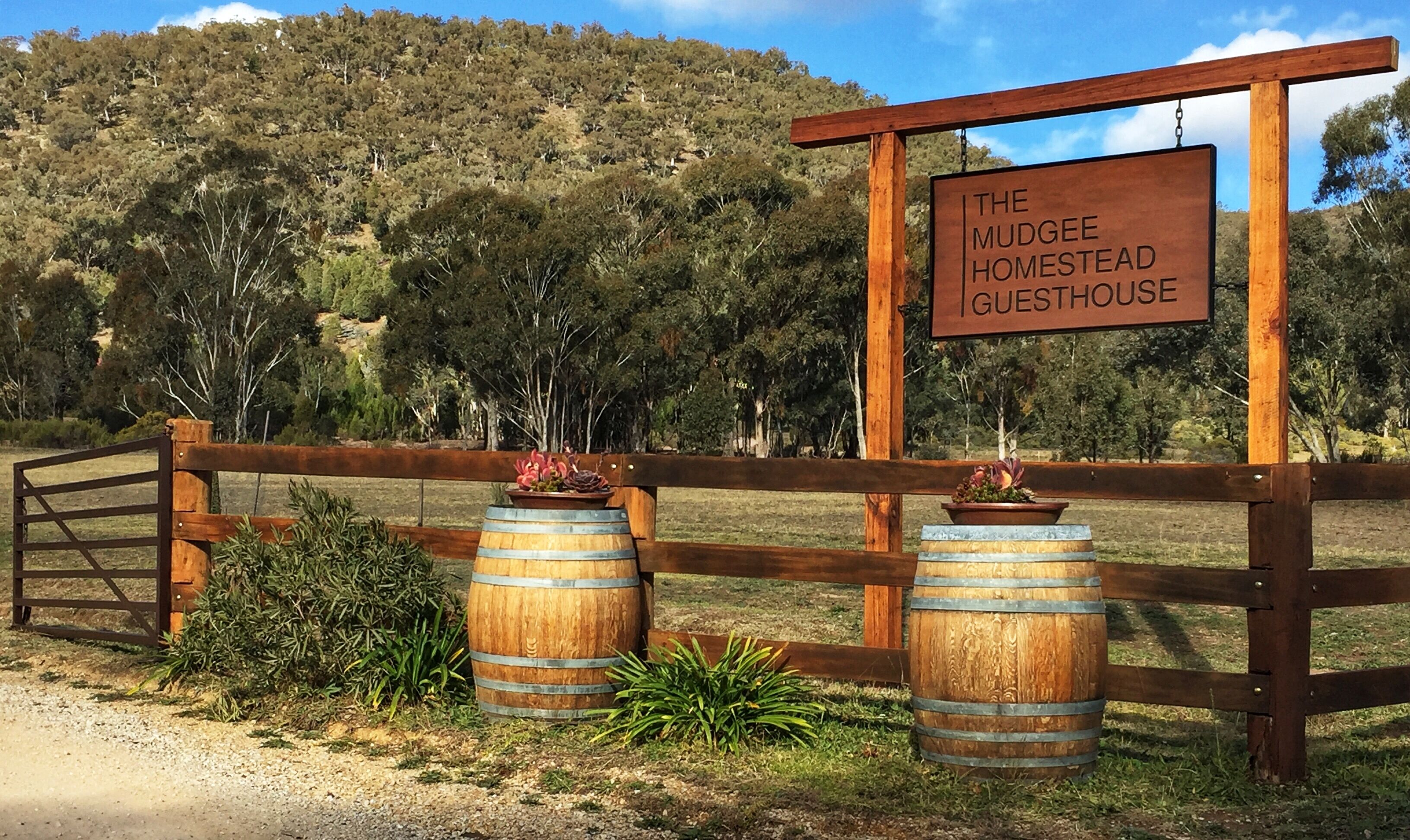Un panneau en bois portant l’inscription « The Mudgee Homestead Guesthouse » et une clôture en bois avec deux barils.